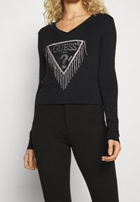 Musta pitkähihainen paita V-kaula-aukolla, jossa edessä kolmionmuotoinen logo, jossa on strassikoristeita. Pehmeä pinta ja vartalonmyötäinen malli.