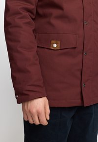 Bordeaux geïsoleerde jas met een gestructureerde afwerking, een borstzak met een bruine leren flap en drukknoop, en een knoopsluiting bij de manchetten.