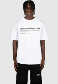 T-shirt en coton blanc avec du texte noir : "la jeunesse est gaspillée sur les jeunes" avec des éléments graphiques. Modèle portant un pantalon cargo noir.