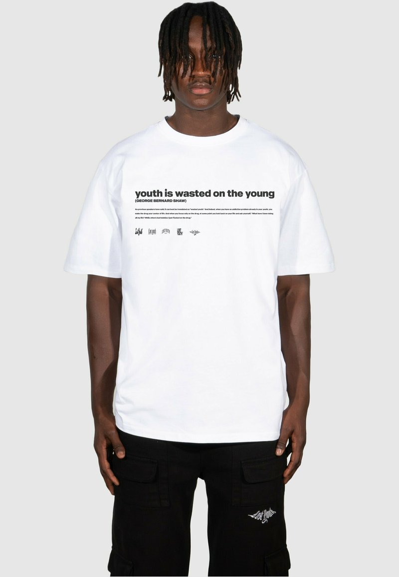 T-shirt en coton blanc avec du texte noir : "la jeunesse est gaspillée sur les jeunes" avec des éléments graphiques. Modèle portant un pantalon cargo noir.