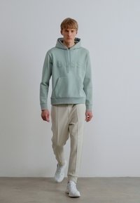 Ανοιχτοπράσινο φούτερ με ανάγλυφο λογότυπο "BOSS", συνδυασμένο με κρεμ χρώματος παντελόνι με στενή γραμμή και λευκά αθλητικά παπούτσια· λείο ύφασμα με τσέπη καγκουρό.