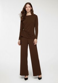Top marrone a maniche lunghe con dettaglio attorcigliato e accentuazione circolare in vita, abbinato a pantaloni marroni a gamba larga. Tessuto di trama liscia.
