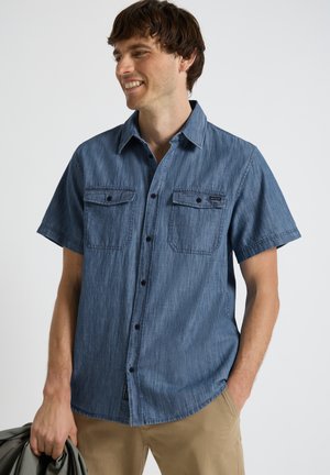 Lachende man met een korte mouwen blauwe spijkerblouse met twee borstzakken, houdt een grijze jas in zijn linkerhand.