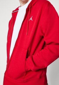 Persoon in een rood zip-up hoodie met een wit Jordan-logo over een wit T-shirt, hand in de voorzak, tegen een effen achtergrond.