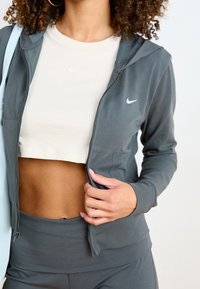 Sudadera gris con cremallera y capucha, con un pequeño logo blanco de Nike en el pecho. Lleva una camiseta corta beige claro debajo; textura suave.