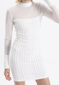 Robe blanche présentant un motif en maille texturée, des manches longues en tissu transparent, un col montant et une silhouette moulante.