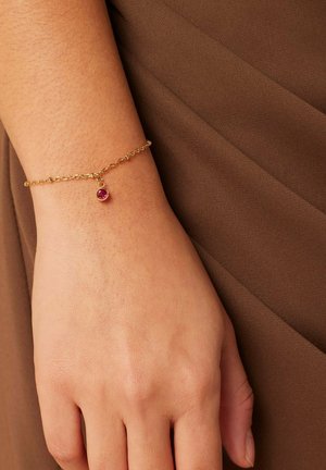 Gouden kettingarmband met een kleine rode edelsteencharme, met een delicaat schakeldesign, op een hand tegen een bruine stoffen achtergrond.