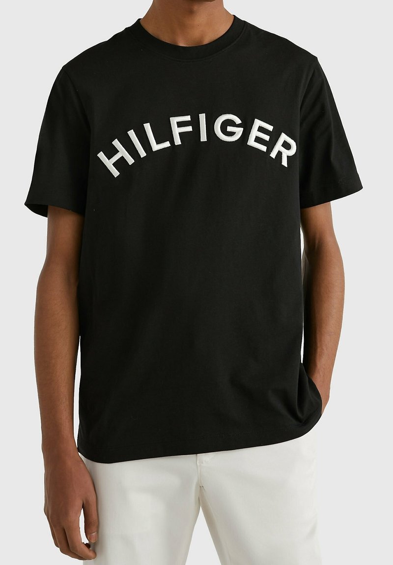 Black cotton t-shirt featuring raised white embroidered "HILFIGER" text in an arch design. Short sleeves and round neckline.