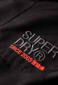 La tela negra presenta el bordado "SUPERDRY" en gris, con "SINCE 2003" en rojo. El hilo contrasta con la textura suave del material.
