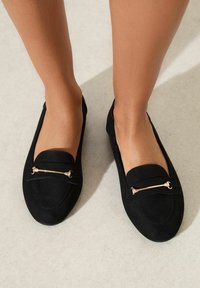 Černé semišové loafers se zlatým ozdobným přezkou. Plochý design s kulatou špičkou a prošitými detaily na nártu.