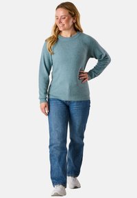 Hellblauer, strukturierten Strickpullover mit Rundhalsausschnitt und gerippten Bündchen, kombiniert mit blauen Jeans und weißen Sneakern. Modell steht.