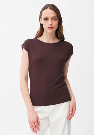 Kobieta ubrana w krótki rękaw ciemnobrązowej bluzki z kontrastową lamówką, w zestawie z białymi spodniami, stojąca na jednolitym białym tle.