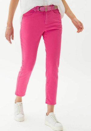 Personne portant un pantalon slim rose vif avec une ceinture tissée assortie, des baskets blanches et un haut blanc ample sur un fond blanc.