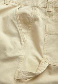 Beige puuvillahousut, joissa on etutasku, sivureikä, näkyvät saumat ja sileä pinta.
