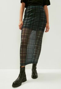 Jupe plissée transparente dans des tons sombres, dotée d'une taille ajustée et de coutures latérales avec des accents verts. Le mannequin est associé à des bottines noires.