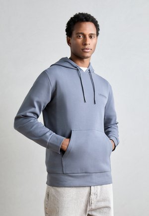 Grijze hoodie met een trekkoord, een voorzak en een geborduurd logo. Zachte stof, normale pasvorm. Gecombineerd met lichte jeans.