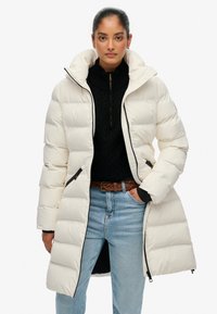 Gewatteerde jas in off-white met een hoge kraag, ritssluiting en twee zijzakken. Getextureerd, doorgestikt ontwerp met zwarte accenten.