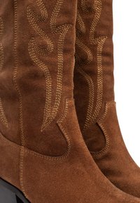 Botas de ante marrón con una textura suave, que presentan un pespunte decorativo de color marrón claro en un patrón curvado. Los tacones gruesos aumentan la estabilidad.