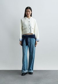 Kvinne iført en hvit cropped jakke med knapper, vide blå jeans med hvite prikker og en marineblå genser knyttet rundt livet.