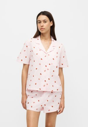 Jonge vrouw draagt een lichtroze, knoopjeshemd met korte mouwen en bijpassende korte broek met kleine rode aardbeiprint, staand tegen een effen witte achtergrond.