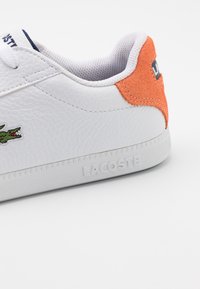 Baskets en cuir blanc avec un patch talon en daim orange, semelle texturée embossée du mot "Lacoste", et logo crocodile vert brodé.