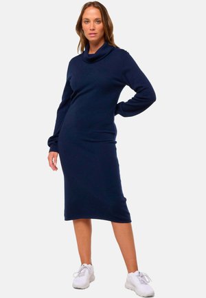Robe en maille bleu marine avec un col drapé, des manches longues et une silhouette ajustée, arrivant mi-mollet, assortie à des baskets blanches.