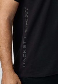 Záber zblízka na ruku osoby, ktorá má na sebe čierne tričko s vertikálnym nápisom "HACKETT SPORT" pozdĺž boku.