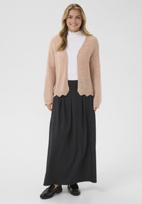 Cárdigan de punto rosa claro, con mangas largas y dobladillo festoneado, superpuesto sobre un suéter de cuello alto blanco, combinado con una falda larga negra y zapatos negros.