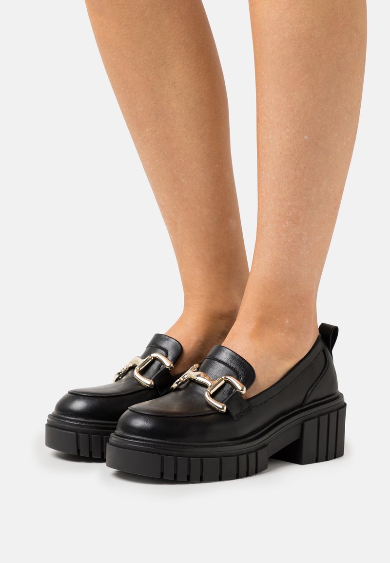 Mocassins noirs en cuir avec une semelle plateforme épaisse et texturée, ornés d'accessoires dorés. Design sans lacets avec un bout rond et une languette à l'arrière.