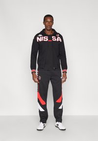 Svart sportjacka med röda bokstäver och logotyp, kombinerad med svarta och röda träningsbyxor med vita detaljer och sidrand.