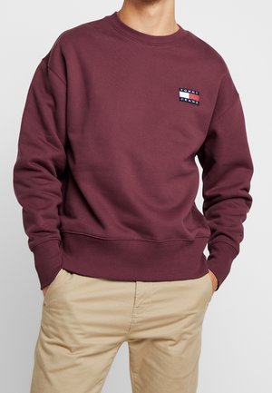 Homme portant un sweat-shirt bordeaux à col rond avec un petit logo Tommy Jeans sur la poitrine et un pantalon beige, les mains dans les poches.