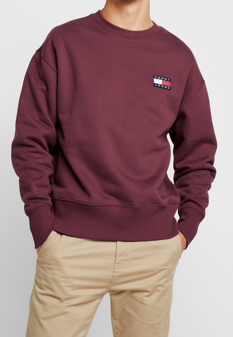 Homme portant un sweat-shirt bordeaux à col rond avec un petit logo Tommy Jeans sur la poitrine et un pantalon beige, les mains dans les poches.