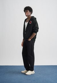 Μαύρο φούτερ με φερμουάρ και λογότυπο, λευκή φανέλα, μαύρο παντελόνι και μπεζ αθλητικά παπούτσια, σε μοντέλο που στέκεται σε μπλε χαλί σε απλό φόντο.