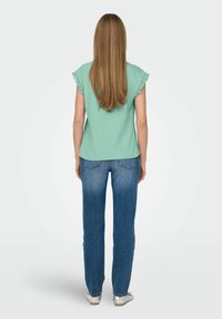 Mintgrünes kurzärmeliges Oberteil mit Spitzenbesatz, kombiniert mit blauen Denim-Jeans. Das Haar des Models ist lang und gerade, von hinten sichtbar.