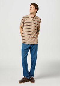 Beige pikétröja med horisontella röda och marinblå ränder, korta ärmar och krage; ihop med blå denimbyxor och bruna seglarskor.