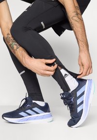Baskets de sport navy avec des rayures bleu clair, semelle blanche et tissu texturé. Leggings noirs ajustés avec détail zippé à la cheville.