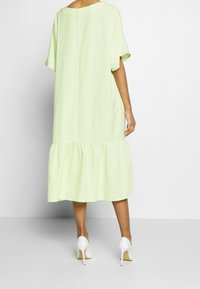 Donna che indossa un vestito midi leggero di colore verde chiaro con maniche corte e orlo arricciato, abbinato a scarpe bianche con tacco alto, in piedi davanti a uno sfondo semplice.