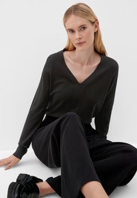 Schwarzer V-Ausschnitt-Pullover mit langen Ärmeln, der eine glatte Textur und Nahtdetails aufweist, kombiniert mit weiten schwarzen Hosen und klobigen schwarzen Schuhen.