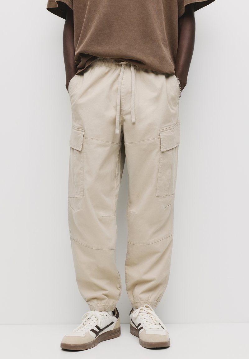 Beige cargobroek met elastische boorden en tailleband. Beschikt over twee zijzakken en twee cargozakken, gemaakt van lichtgewicht stof.