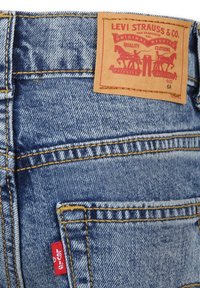 Jeans de mezclilla Levi's con tejido azul, costuras amarillas y una etiqueta de cuero marrón con un logotipo y diseño rojo en la cinturilla trasera.