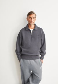 Donkergrijze ritstrui met een opstaande kraag en geribbelde manchetten, in combinatie met grijze joggingsbroek met een accentdetail op het been.