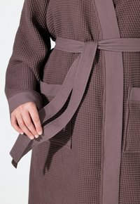 Peignoir en maille waffle dans un mauve doux, avec des manches longues, une ceinture à la taille, de grandes poches et des détails en tissu texturé.