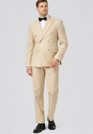 Homme debout portant un costume beige croisé à boutons dorés, une chemise blanche, un nœud papillon noir et des chaussures habillées noires sur un fond uni.