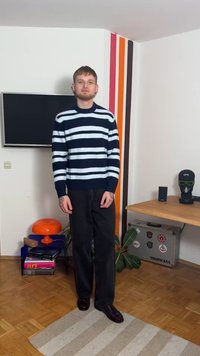 Marineblauer und weiß gestreifter Strickpullover mit hohem Kragen, kombiniert mit schwarzen, locker sitzenden Hosen und dunklen Schuhen, stehend vor einer weißen Wand.