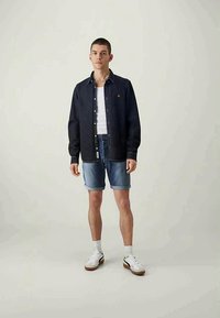 Camisa de ganga escura com botões sobre uma t-shirt branca, combinada com calções de ganga azul-claro e ténis brancos com solas castanhas.