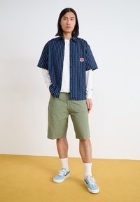 Navy short-sleeve shirt with white vertical stripes, white long-sleeve shirt underneath, green shorts, and light blue sneakers.