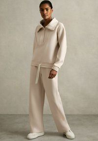 Helle beige Zip-up Sweatshirt mit hohem Kragen und passender Hose. Weicher Stoff mit lockerer Passform, ausgestattet mit einem Kordelzugbund und weitem Bein.