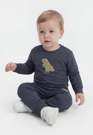 Jeune enfant aux cheveux clairs portant un sweat-shirt gris foncé à motif dinosaure et un pantalon, assis en tailleur sur un sol blanc avec des baskets blanches.