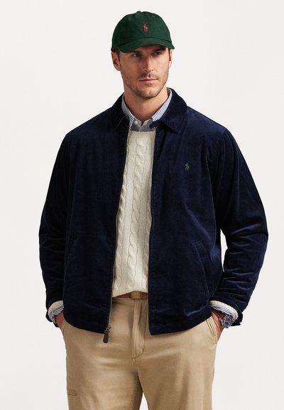 Navy corduroy jacket with a pointed collar, green embroidered logo, paired with a cream cable-knit sweater and tan trousers.