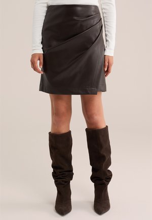 Donna che indossa una gonna in pelle marrone scuro e stivali alla nappa morbidi marrone scuro al ginocchio, con gambe nude e una maglia bianca a maniche lunghe.